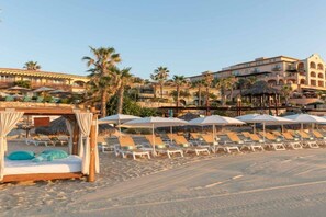 A beachside hotel with sun loungers and umbrellas, a cabana, and a building with arches in the background.