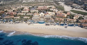 An aerial view of a coastal resort with multiple buildings, a sandy beach, and several swimming pools.