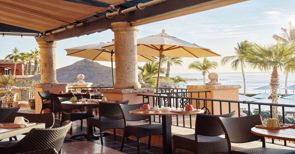 Un área de estar al aire libre con mesas, sillas, sombrillas y vista a la playa y al océano.