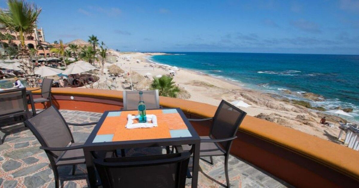 Terraza frente al mar con una mesa y sillas, con vista a una playa y al océano.