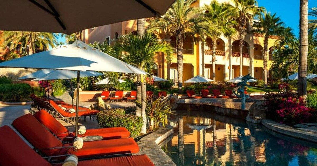 A poolside area with lounge chairs and umbrellas, surrounded by palm trees and a building with arches.