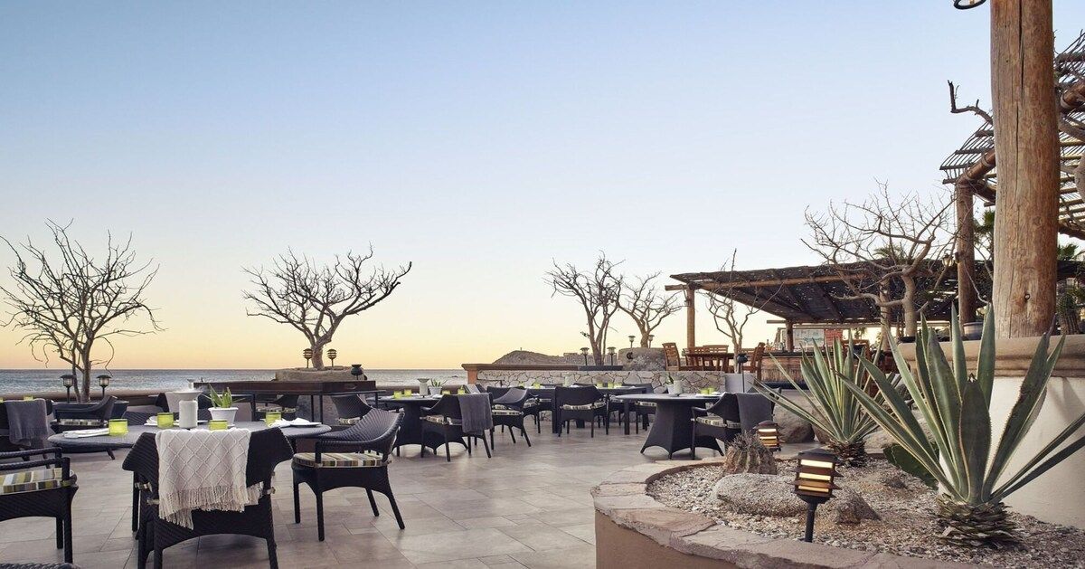 An outdoor dining area with tables and chairs, a view of the sea, and a building with a traditional architectural style.