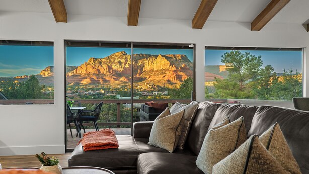 Un salon moderne avec un canapé en cuir, de grandes fenêtres donnant sur une vue sur les montagnes et un balcon aménagé avec du mobilier d’extérieur.