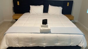 Un lit bien fait, avec une tête de lit sombre, du linge de lit blanc et une pile de serviettes pliées au pied du lit.