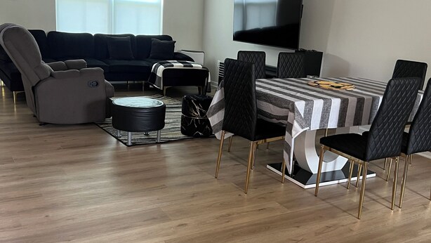 Un salon moderne avec un canapé de couleur foncée, une table à manger avec des chaises, un téléviseur à écran plat et une grande fenêtre équipée de stores.