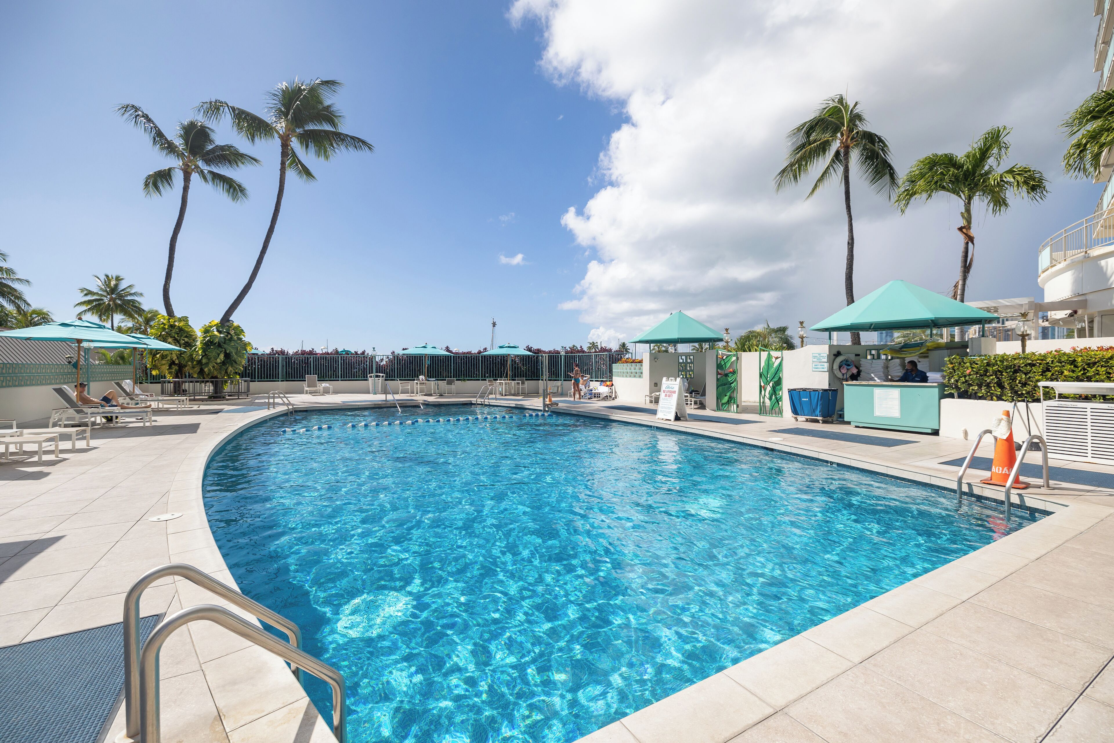 A swimming pool with clear blue water, surrounded by lounge chairs and umbrellas, palm trees, and a small gazebo.