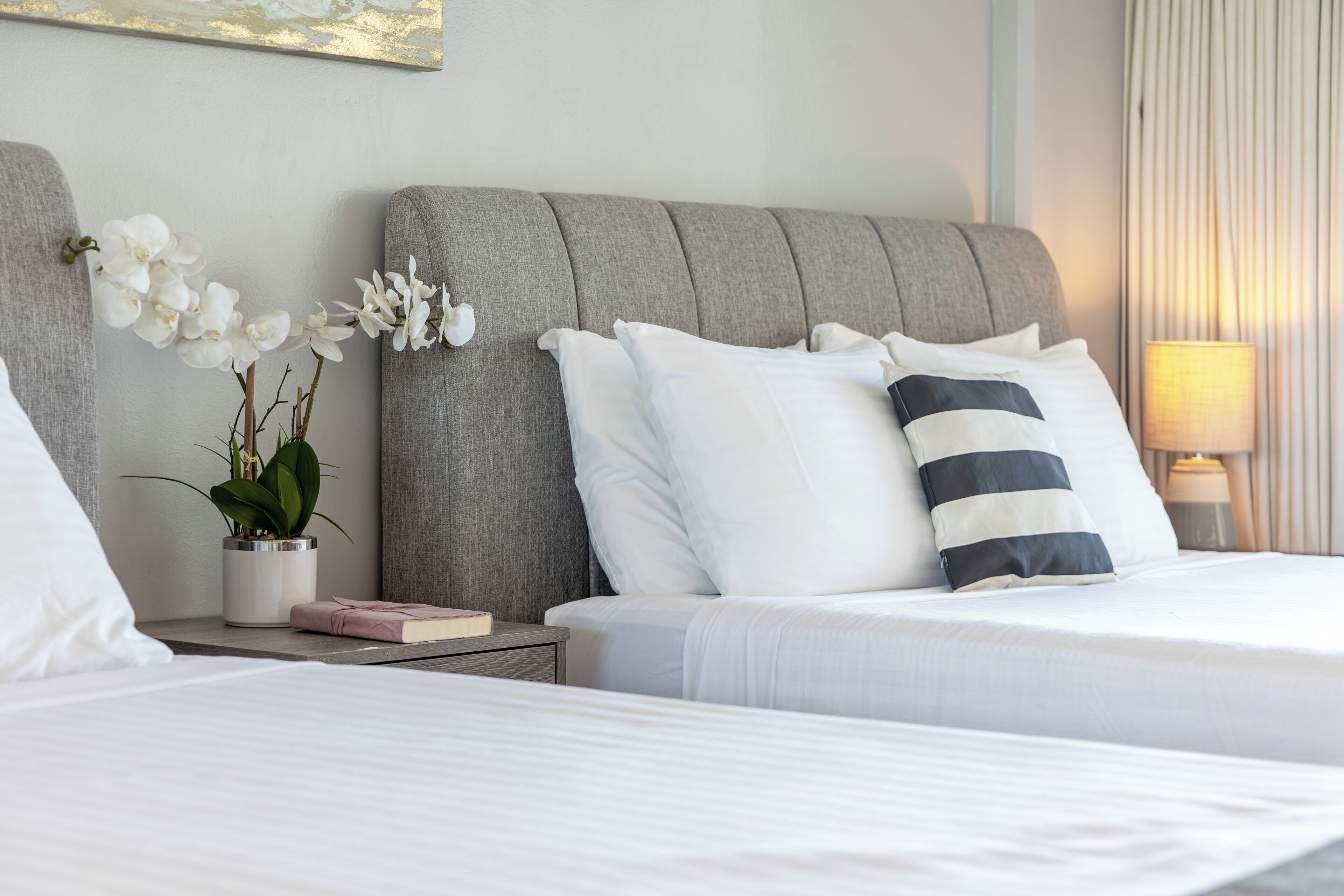 Een net opgemaakt bed met witte en gestreepte kussens, een grijs gestoffeerd hoofdbord, een nachtkastje met een plant en een boek, en een lamp aan de rechterkant.