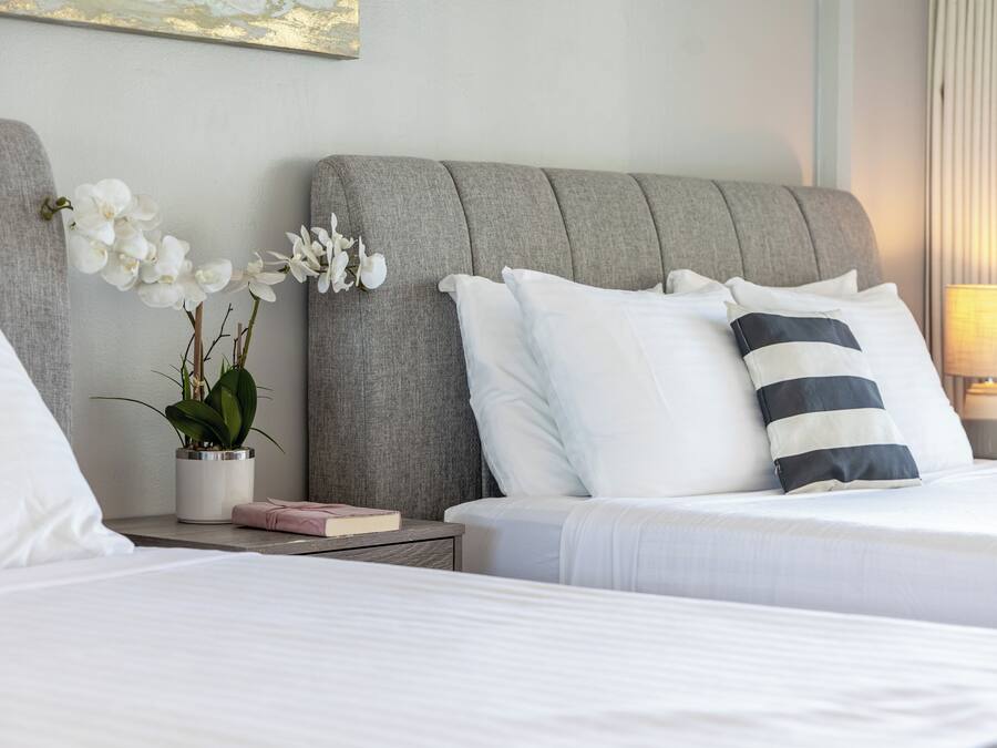 Een net opgemaakt bed met witte en gestreepte kussens, een grijs gestoffeerd hoofdbord, een nachtkastje met een plant en een boek, en een lamp aan de rechterkant.