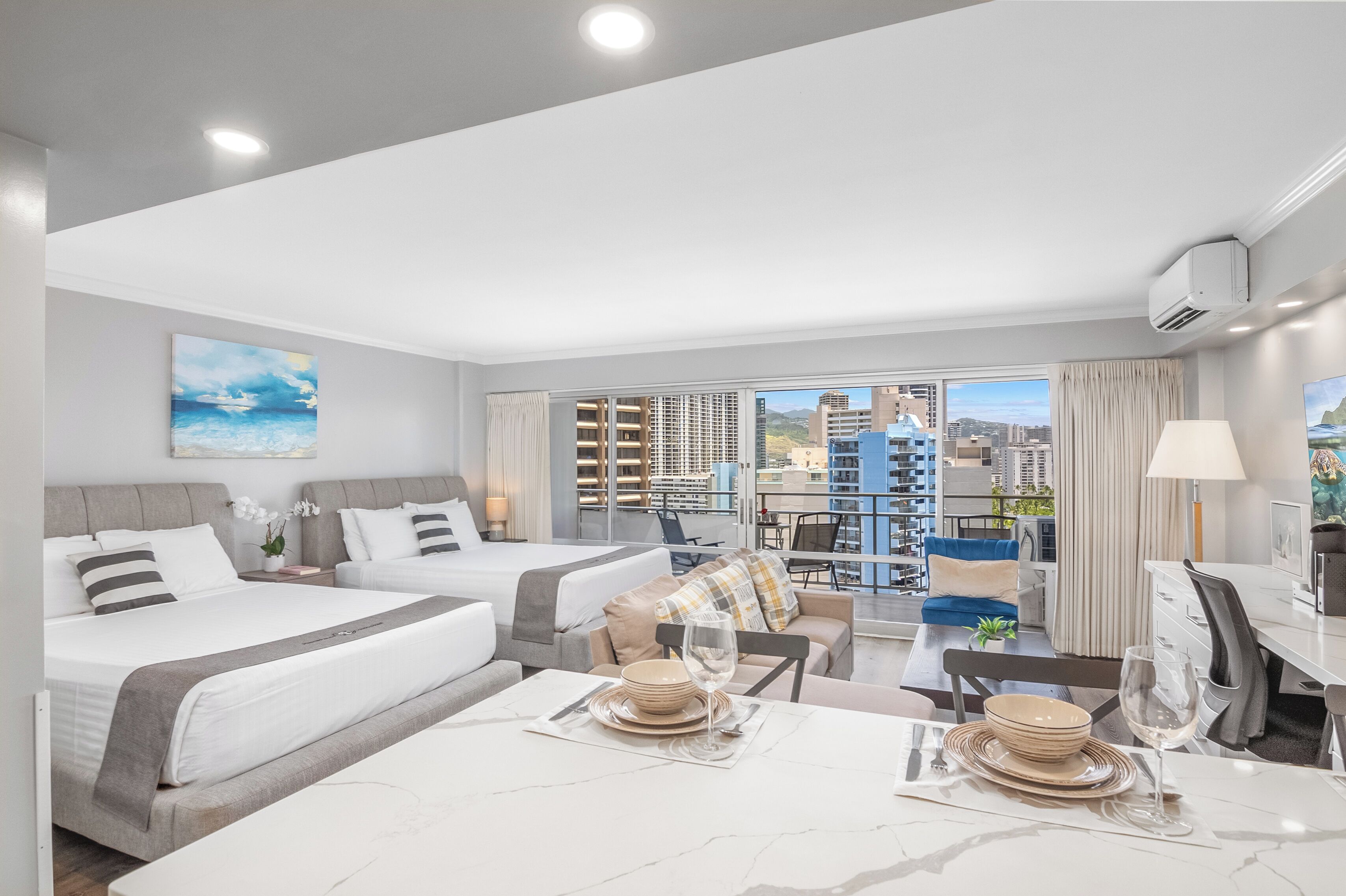 A modern hotel room with two beds, a dining area, and a balcony with a city view.