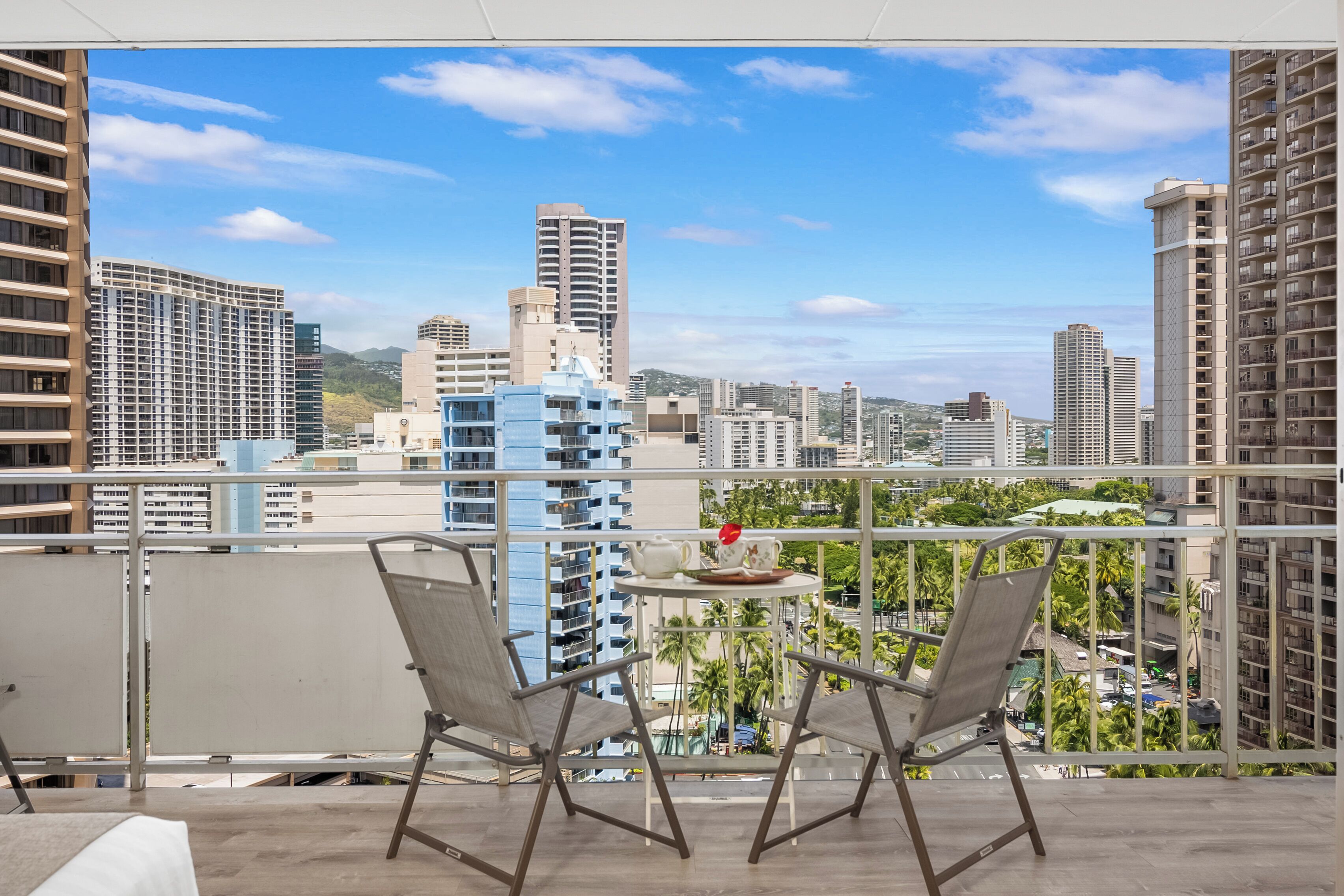 A balcony with two chairs and a table, overlooking a cityscape with various buildings and greenery.