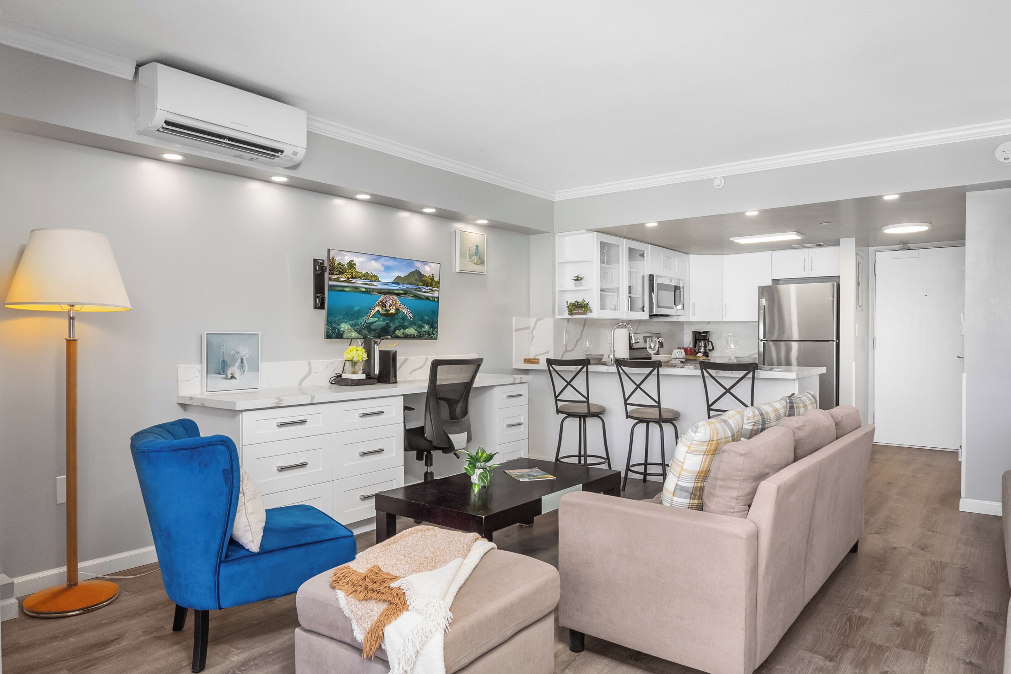 Una sala de estar moderna con un sofá, una silla azul, una mesa de centro y una lámpara. Se observa una zona de cocina con armarios blancos, un refrigerador y un microondas.