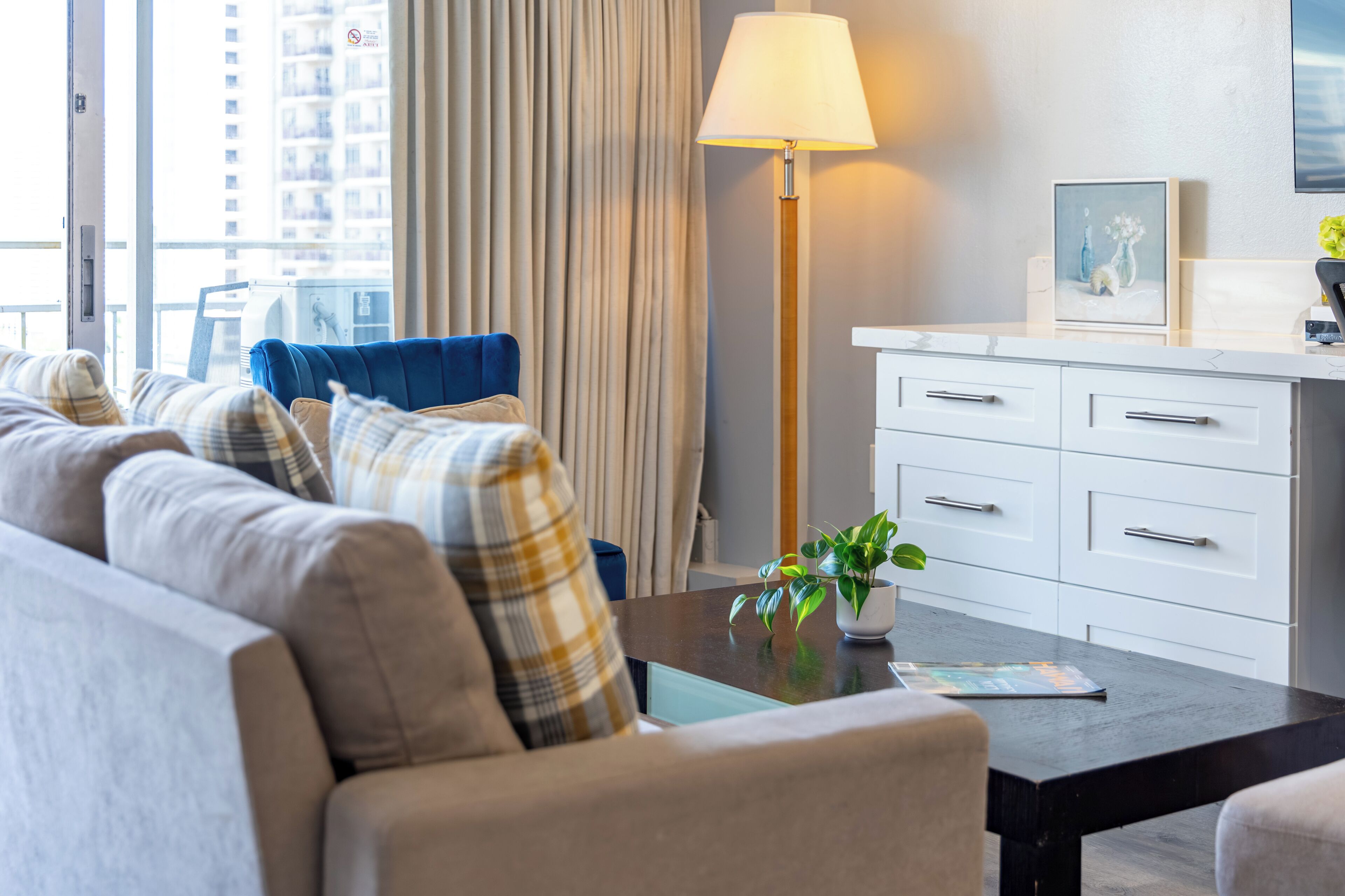 Una habitación de hotel moderna con un sofá, una mesita de centro con una planta, una lámpara y vistas a la ciudad a través de amplios ventanales.