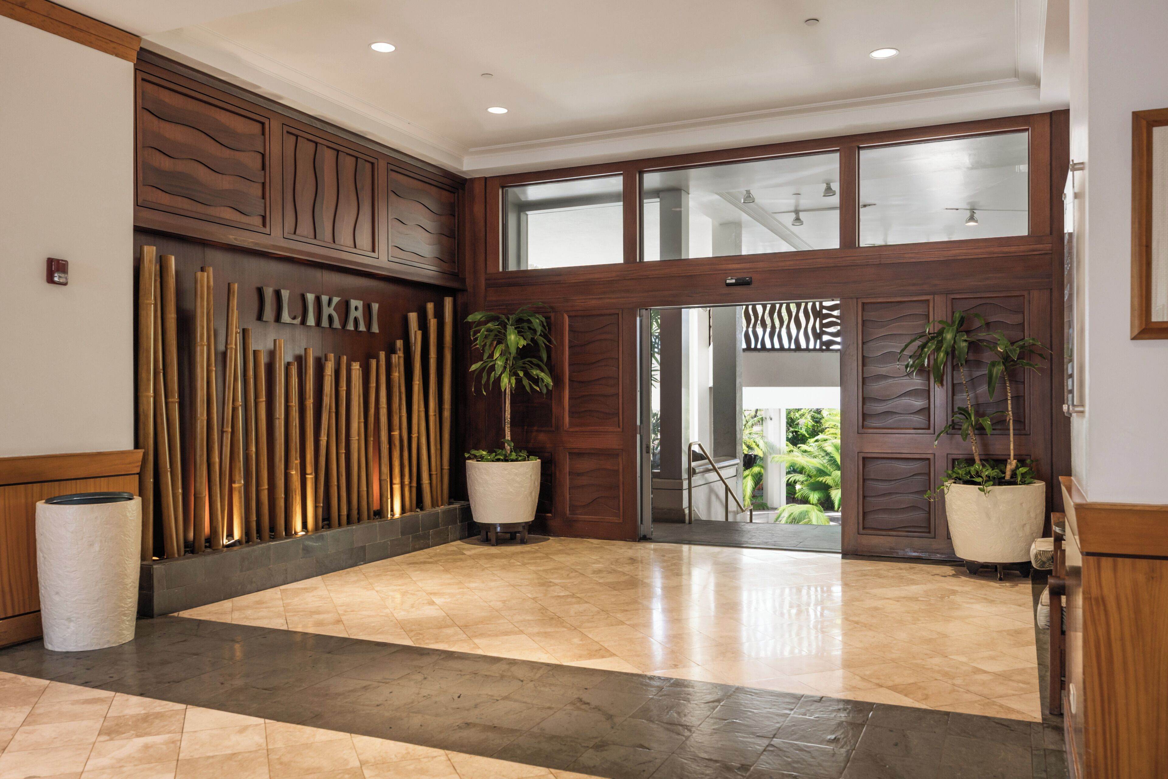 A hotel lobby with a wooden reception desk, a bamboo wall, and a glass door leading to a balcony with plants.
