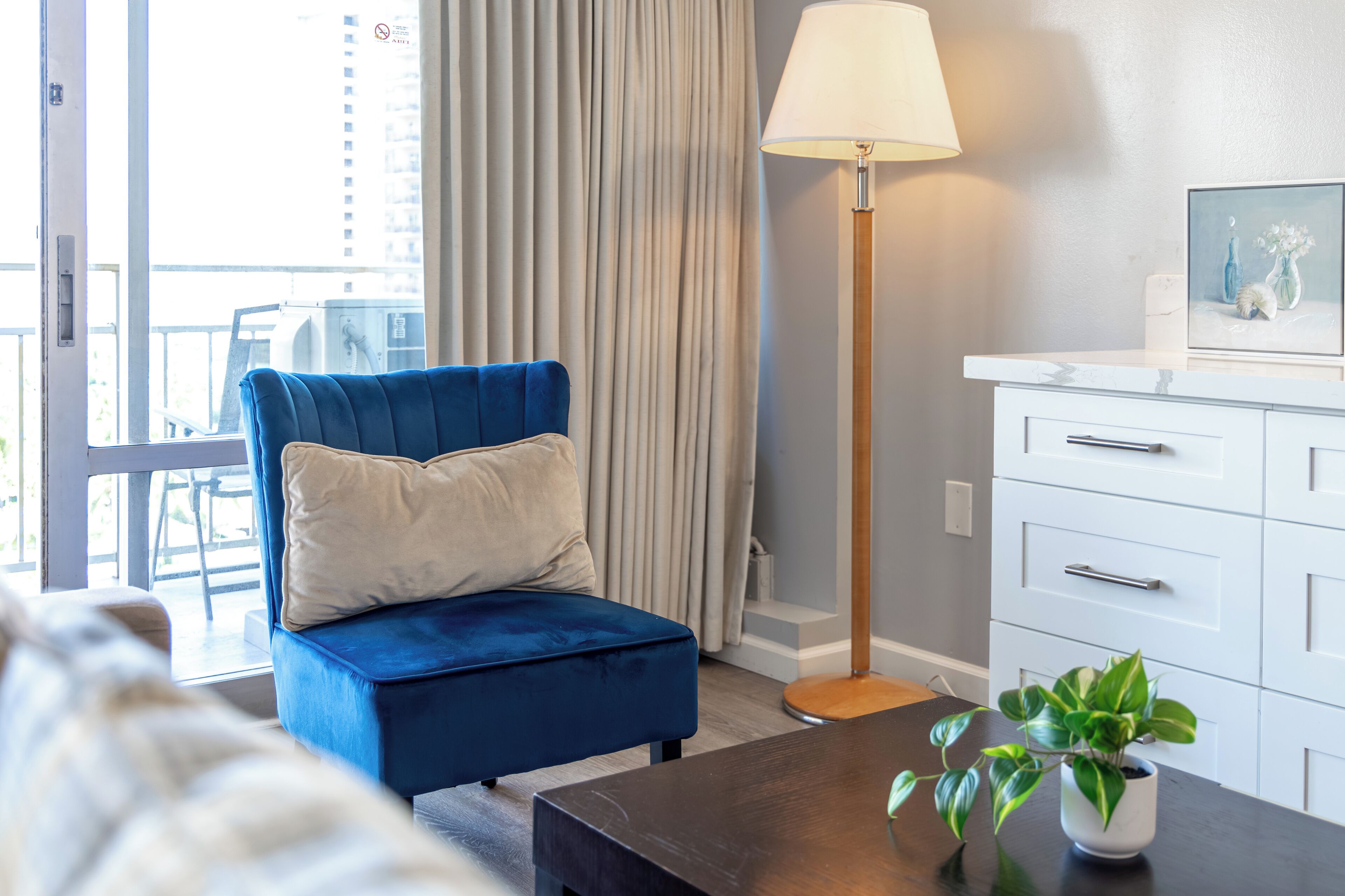 A modern hotel room with a blue armchair, a floor lamp, a small table with a plant, and a white dresser.