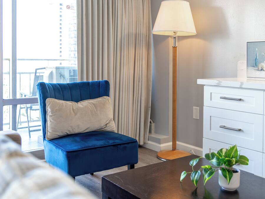 A modern hotel room with a blue armchair, a floor lamp, a small table with a plant, and a white dresser.