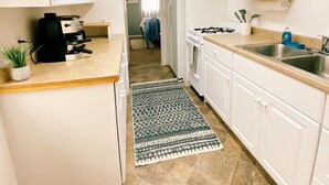 Una cocina estrecha con armarios blancos, una encimera con cafetera, estufa y lavabo. Un tapete estampado cubre el suelo, y se observa un ventilador de techo.