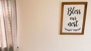 Una mesa de comedor con dos individuales tejidos y una planta pequeña, rodeada de sillas negras. Un cartel enmarcado en la pared dice "Bless our nest".