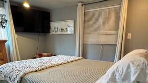 Un dormitorio con cama, ventilador de techo, televisor montado y ventana con persianas.