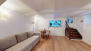A living room with a sofa, a dining table with chairs, a TV mounted on the wall, and a wooden staircase. - Studio Castelnau (Strasbourg)