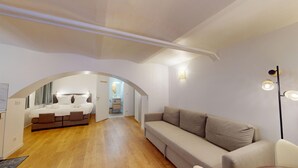 A modern hotel room with a wooden floor, a sofa, a bed with white linens, and a bathroom visible through an arched doorway. - Studio Castelnau (Strasbourg)