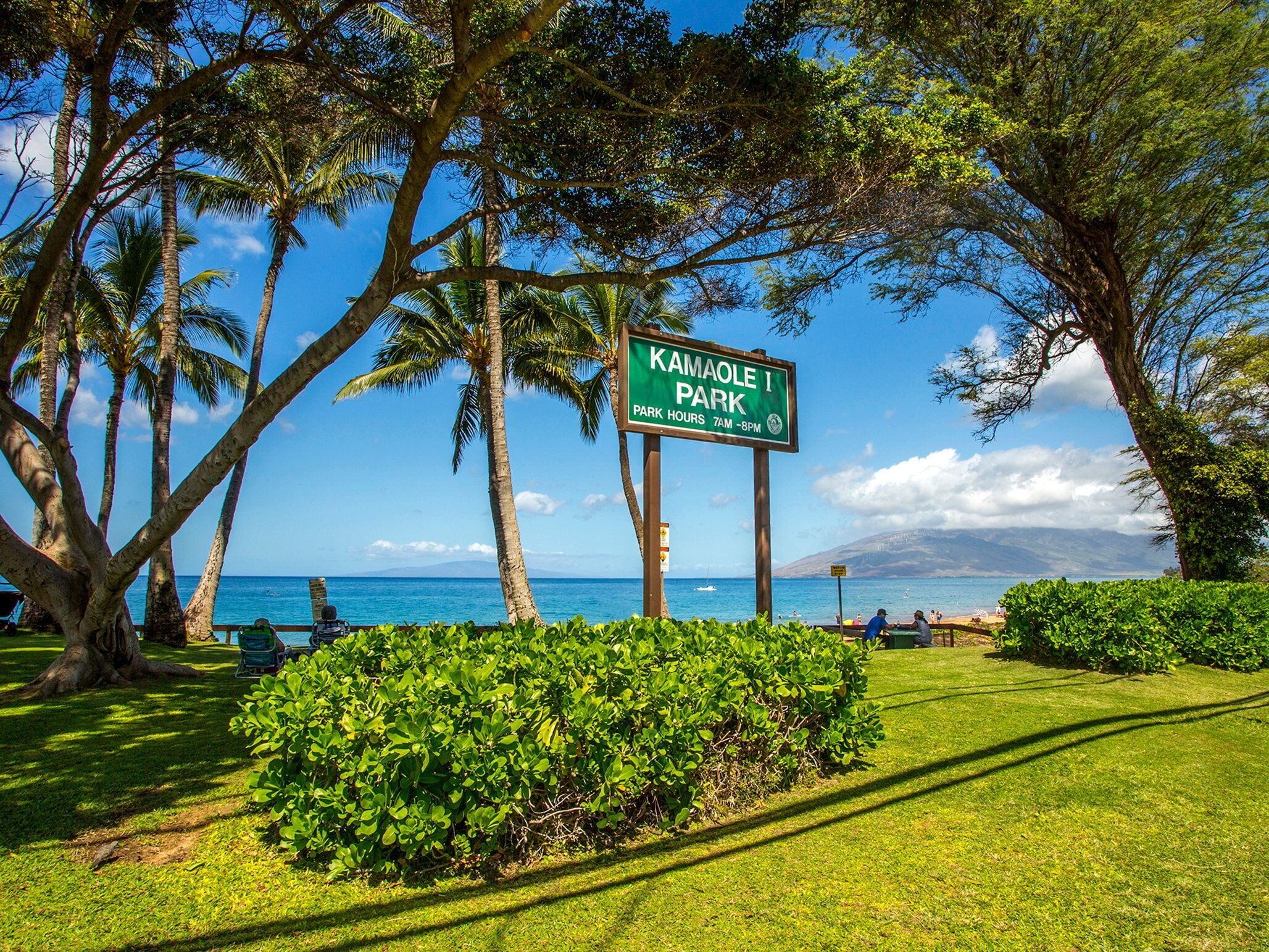 A sign for Kamaole I Park with a view of the ocean and palm trees.