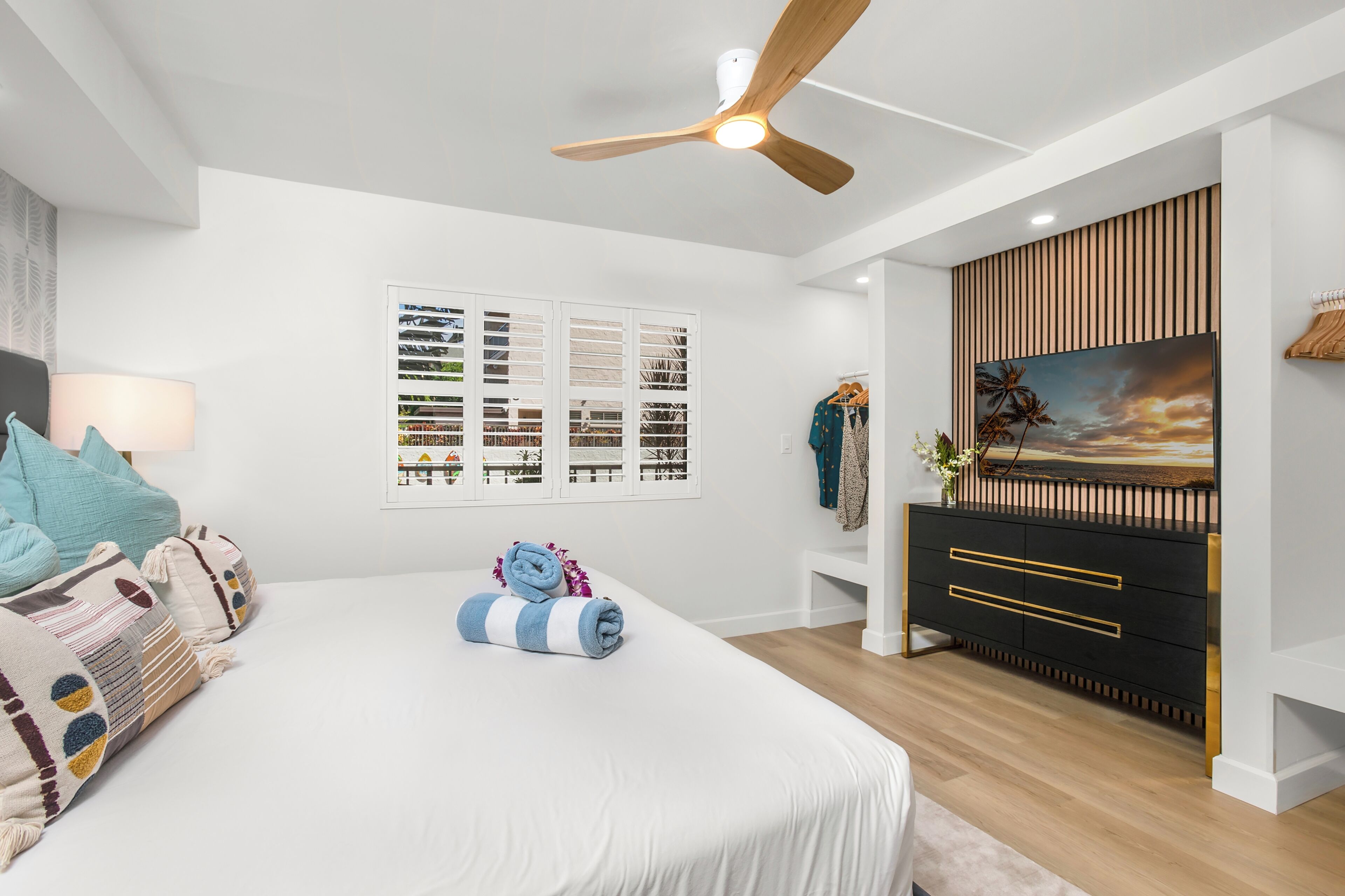 Ein Schlafzimmer mit Bett, Deckenventilator, Wandfernseher, Kommode und Fenster mit Jalousien.