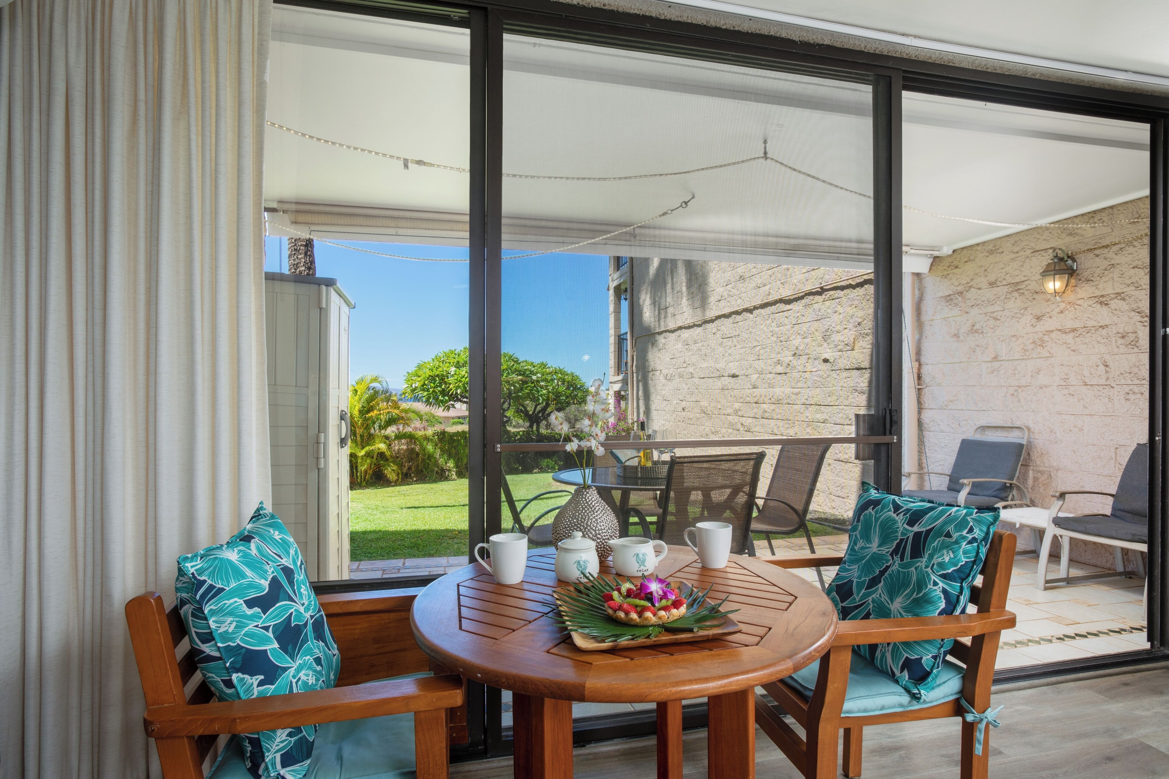 Une table en bois avec des chaises, un parasol blanc et une vue sur un jardin, visible à travers de grandes portes vitrées.