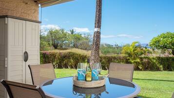 A round glass table with wicker chairs, a tray with two wine glasses and bottles, and a view of a lush garden and palm tree.