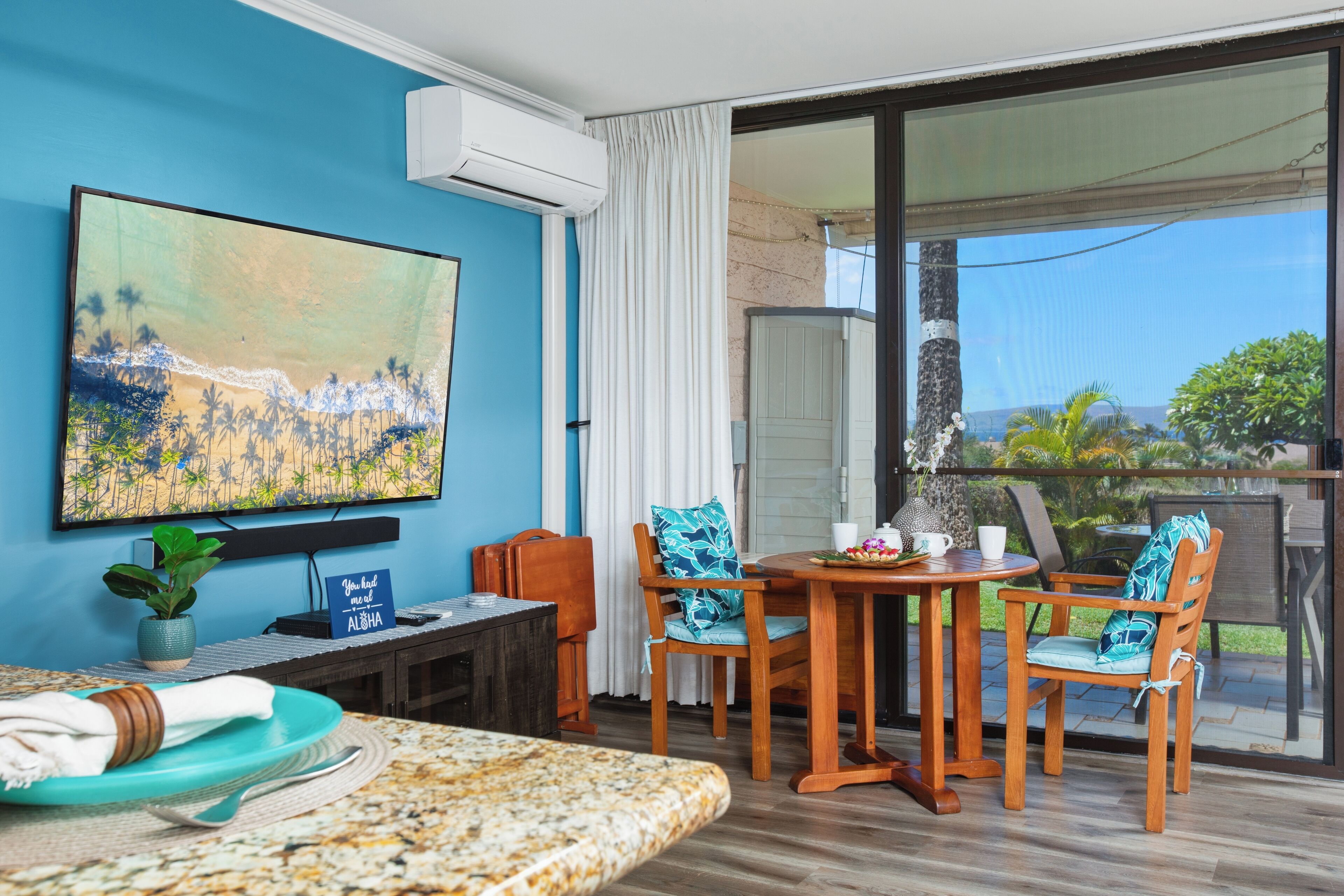 A room with a large painting, a dining table with chairs, a small table with a plant, and a view of the outdoors.