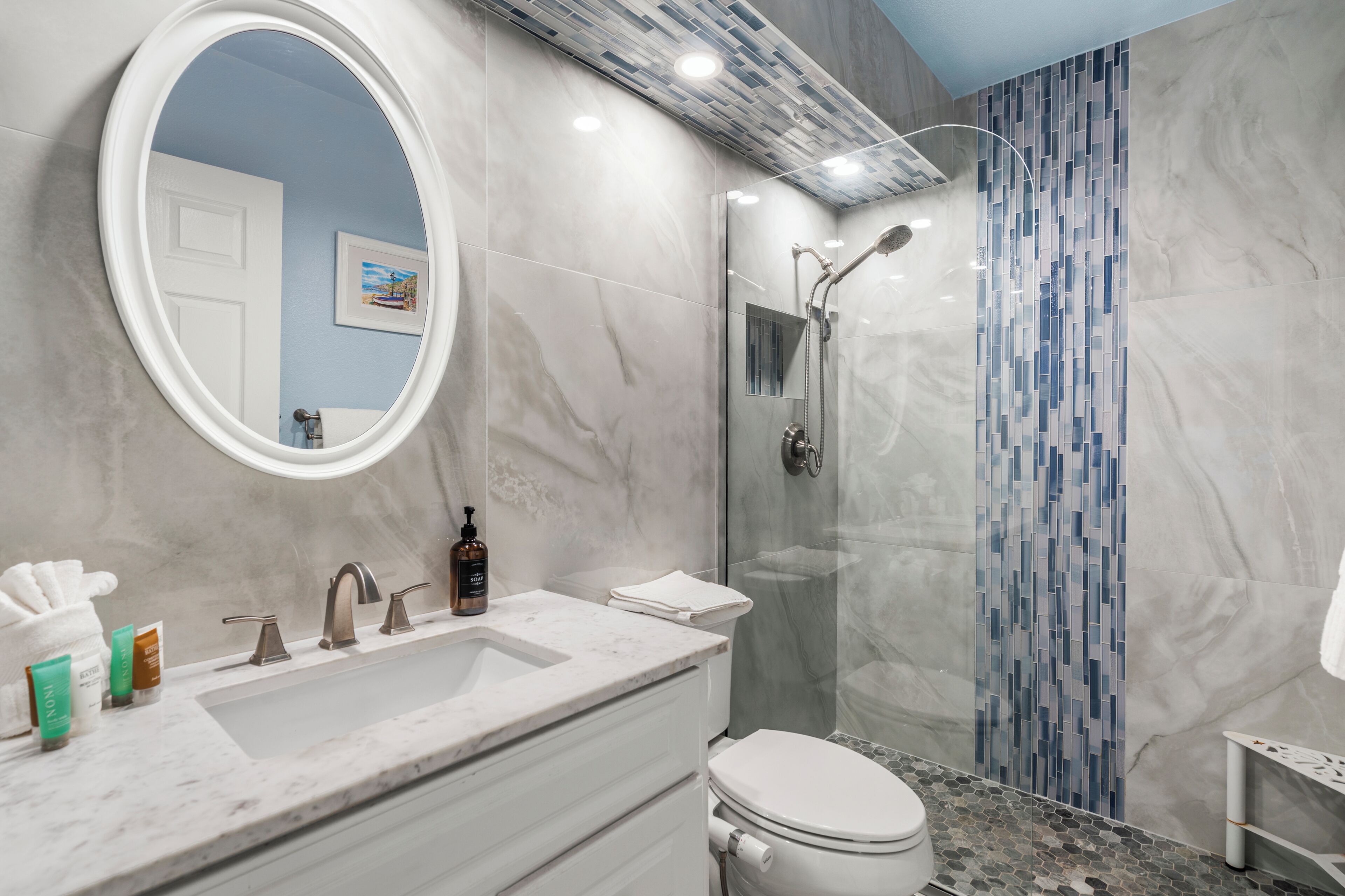 Un baño moderno con un espejo ovalado grande, un lavabo con encimera de mármol, un inodoro y una ducha con mampara de vidrio.