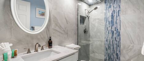 Un baño moderno con un espejo ovalado grande, un lavabo con encimera de mármol, un inodoro y una ducha con mampara de vidrio.