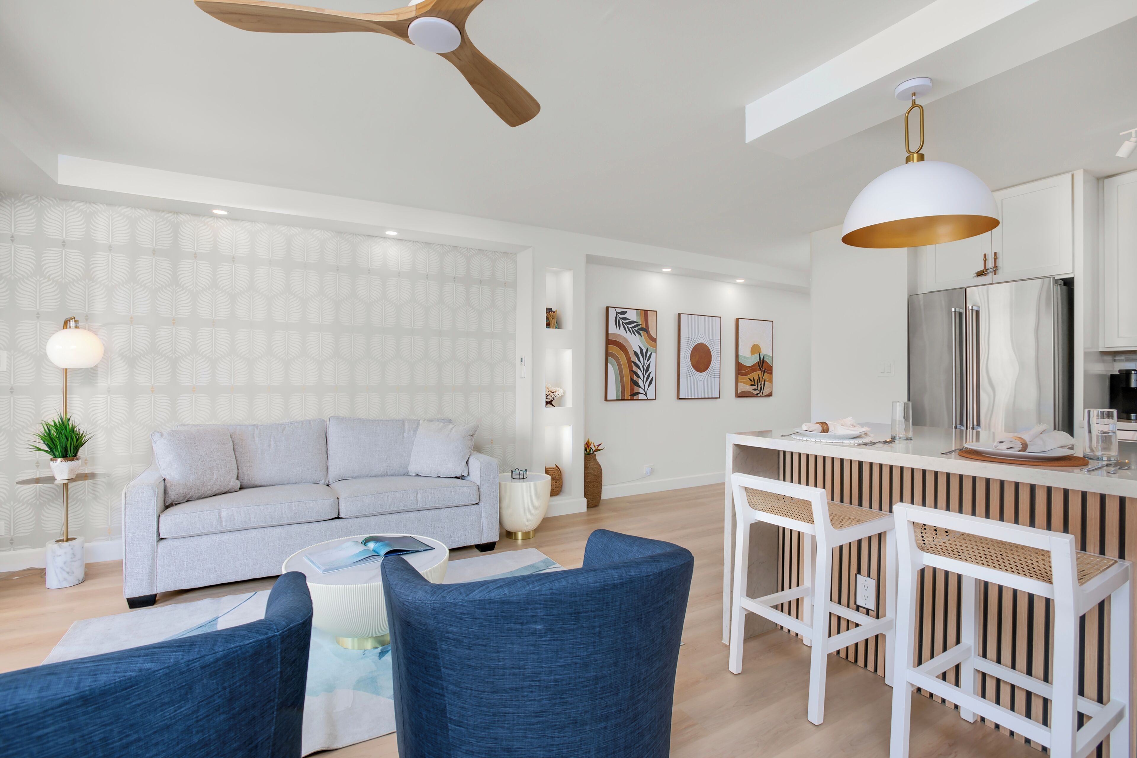 A modern living room with a grey sofa, blue chairs, a dining area with white bar stools, a kitchen with a stainless steel refrigerator, and a ceiling fan.