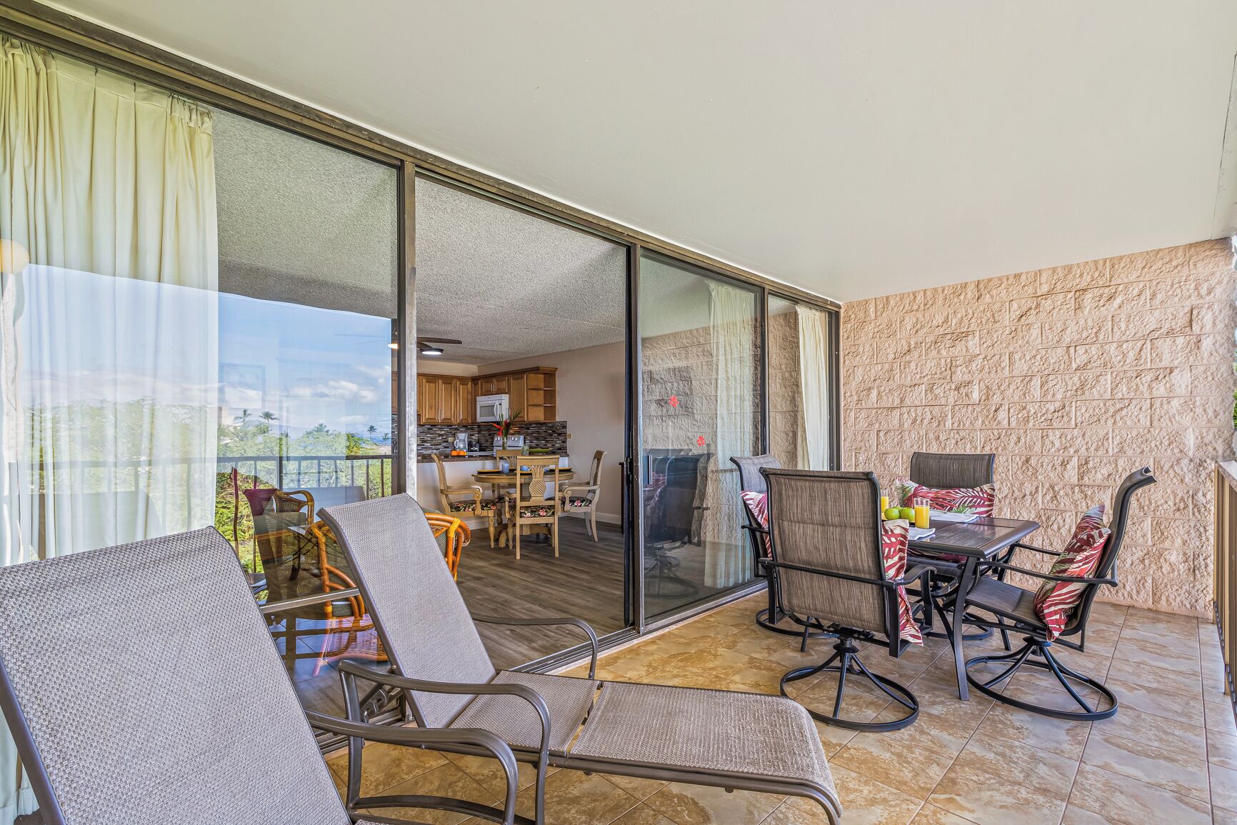 A balcony with wicker chairs and a dining table, a kitchen in the background, and a stone-textured wall.