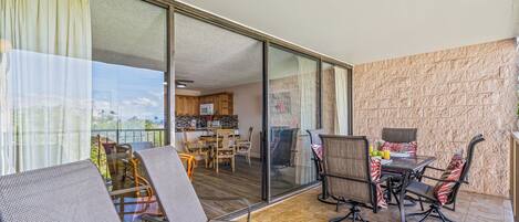 A balcony with wicker chairs and a dining table, a kitchen in the background, and a stone-textured wall.
