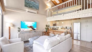 Un vaste espace de vie comprenant un grand téléviseur à écran plat, un ensemble de canapés, un coin repas avec cuisine et un ventilateur de plafond.