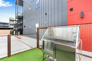 Un espacio exterior moderno con una estación de parrilla metálica, un área de juegos cercada y un edificio con fachada gris y roja.