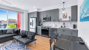 A modern kitchen with grey cabinets, a stainless steel refrigerator, a microwave, and a stove. There is a dining area with a table and chairs, and a large window offering a view of the outside. - Pacific Island Inspired Stay | Amazing Location (New Braunfels)