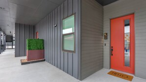 Une entrée moderne avec une porte rouge, un bac à fleurs vert et un paillasson sur lequel est écrit « BONJOUR ».
