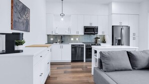 Une cuisine moderne avec des meubles blancs, un four à micro-ondes encastré, un réfrigérateur en acier inoxydable et un coin repas agrémenté d’un canapé gris.