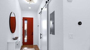 Un couloir avec un mur blanc, une porte rouge, une console blanche sur laquelle est posé un bol, un tapis à motifs et un thermostat.
