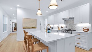 Moderni keittiö, jossa on valkoiset kaapit, suuri keittiösaareke, riippuvalaisimet ja puiset baarituolit.