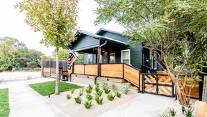 A modern house with a dark exterior, a wooden deck, and a well-maintained garden with small plants and a tree. - Best Location in Downtown NB | Golf Cart Included! (New Braunfels)
