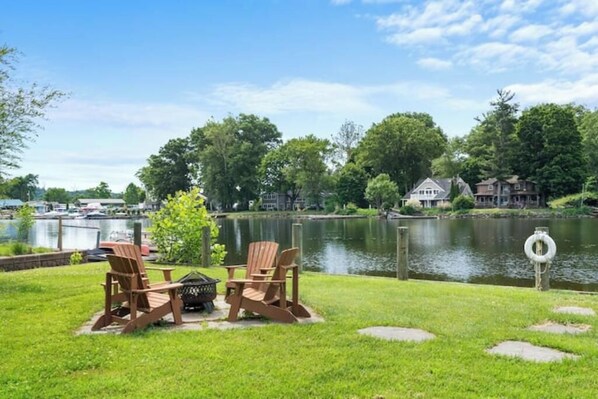 A lakeside patio with Adirondack chairs and a grill, overlooking a calm lake and residential buildings. - Modern Lakefront Retreat (Saugerties)