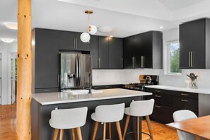 A modern kitchen with dark cabinets, a stainless steel refrigerator, and a central island with bar stools. - Modern Lakefront Retreat (Saugerties)