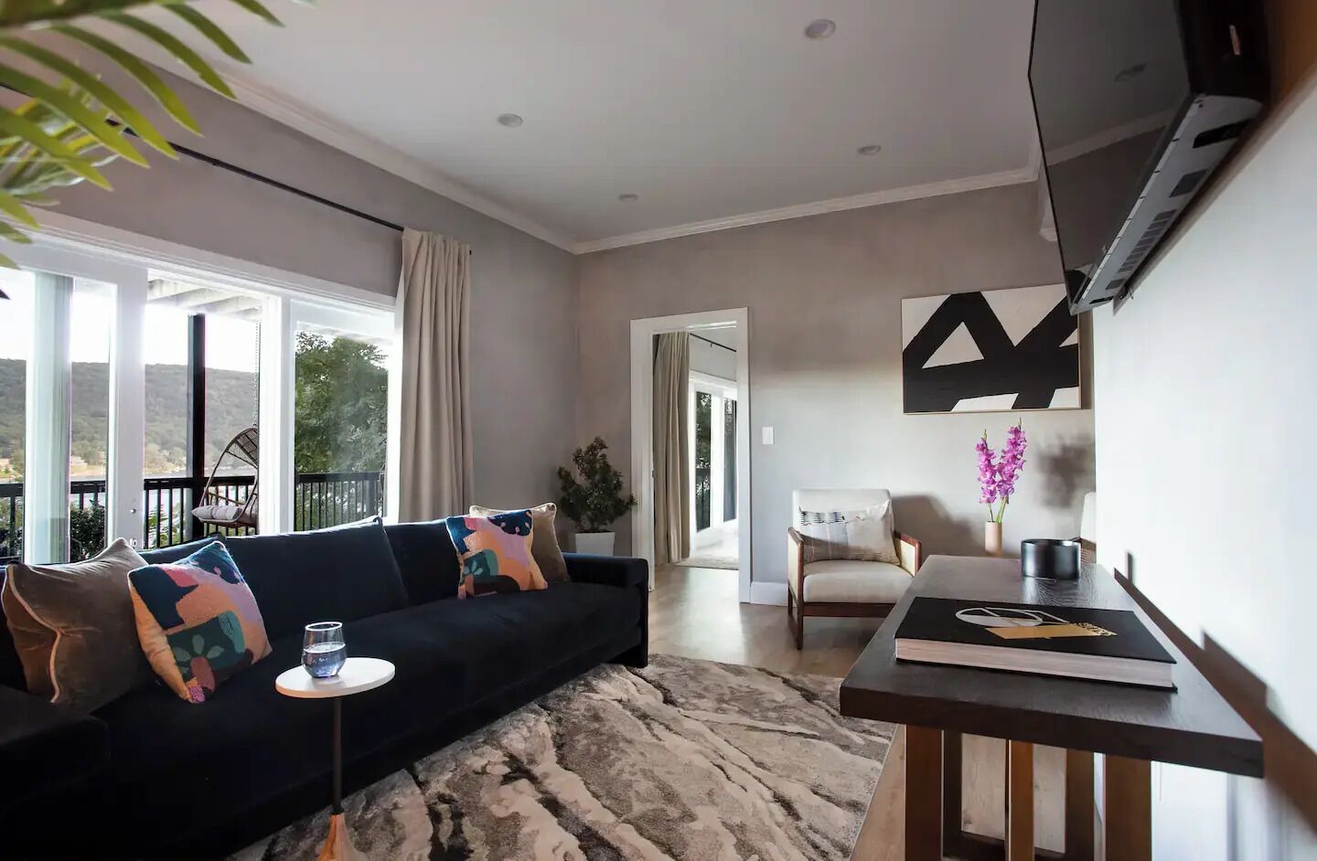 A modern living room with a dark sofa, a coffee table, and a TV. There is a large window with a view of greenery outside.