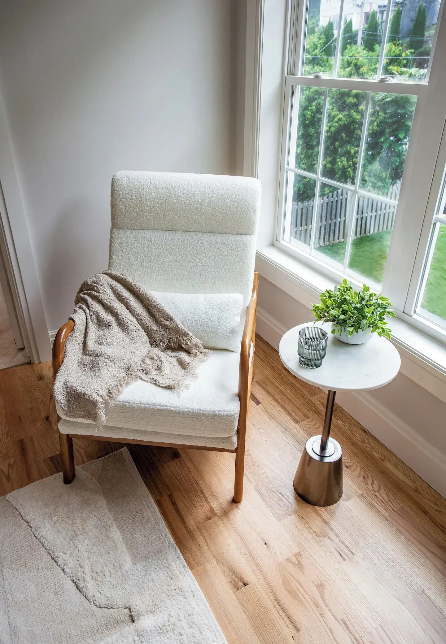 A chair with a white blanket, a small table with a plant, and a window with a view of greenery.