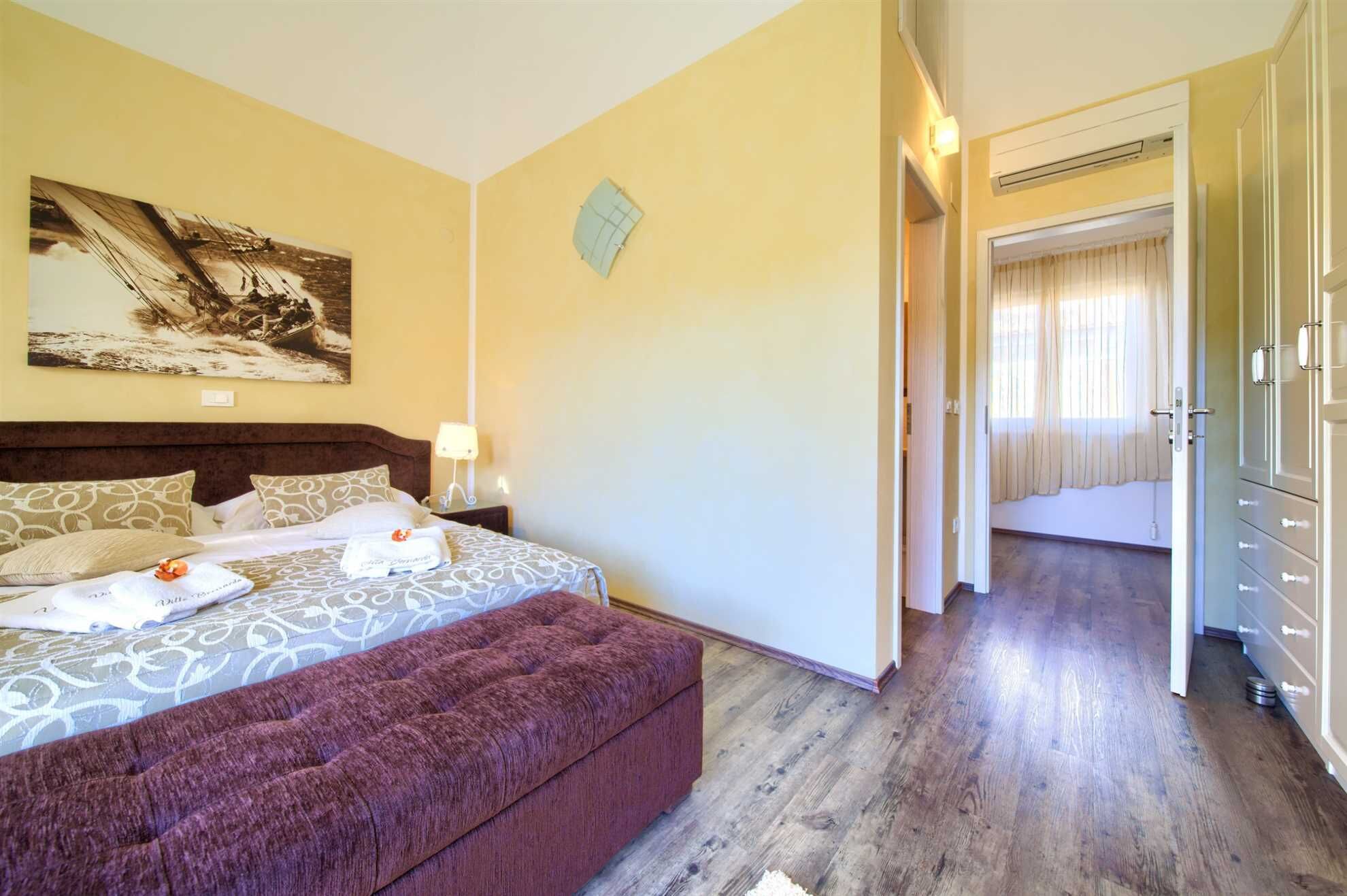 A hotel room with a large bed, a nightstand, a bedside table, a red upholstered bench, a painting on the wall, and a wardrobe.