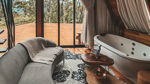 A room with a large window, a sofa, a bathtub, and a wooden table. - Recanto Lavi (Urubici)