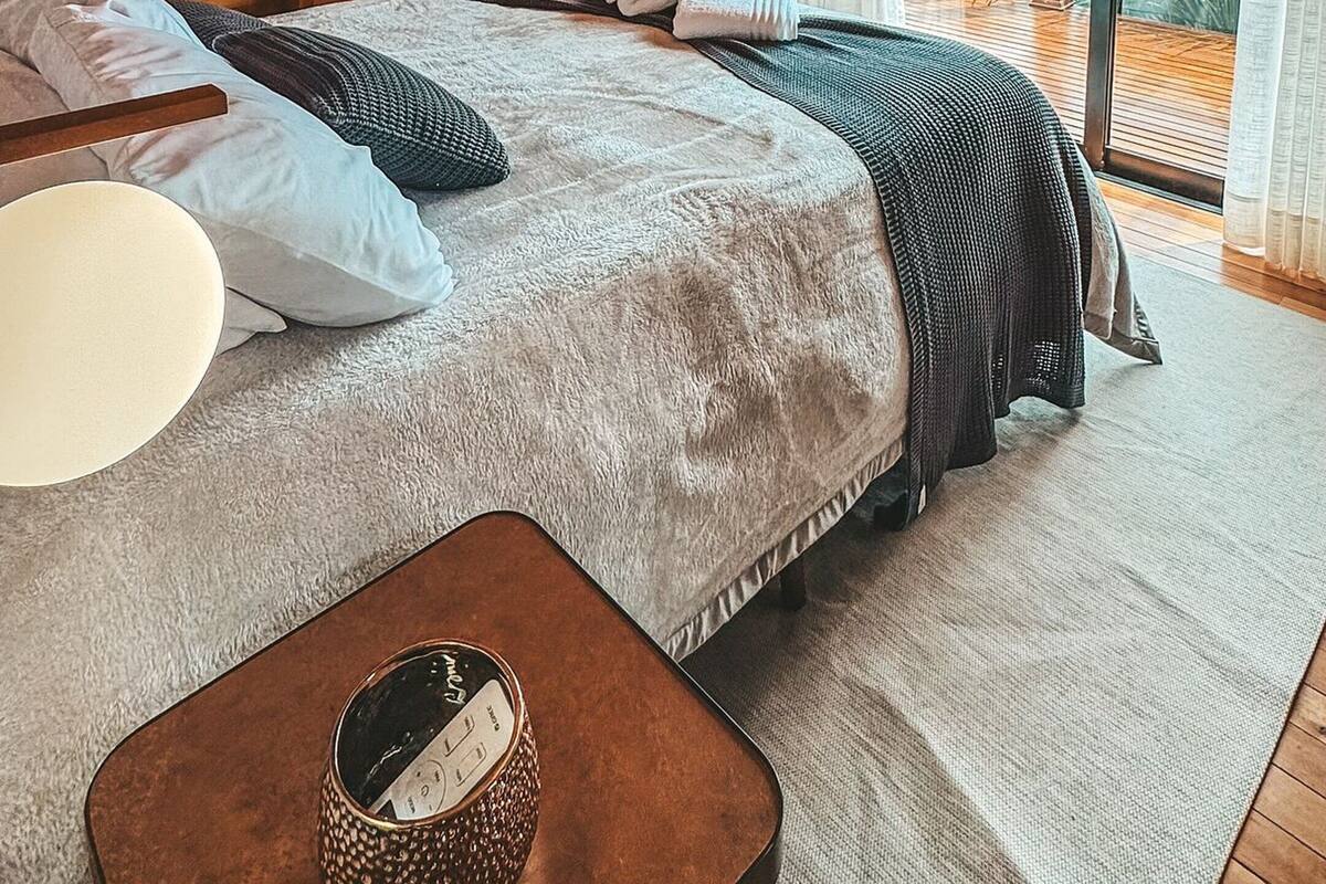 A bedroom with a wooden ceiling, a bed with pillows and a blanket, a bedside table with a watch, and a view of the outdoors.