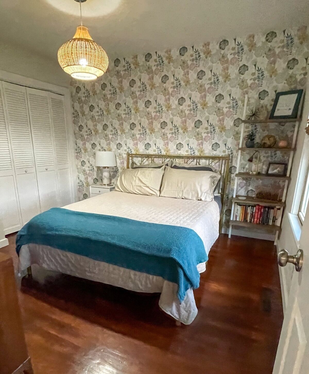 A bedroom with a bed, a wooden floor, a wall with floral wallpaper, a bookshelf, and a hanging light fixture.